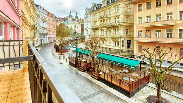 A street view with a row of colorful buildings, a restaurant with outdoor seating, and a tram in the background. - ValenciaKV Apartments (Karlovy Vary)