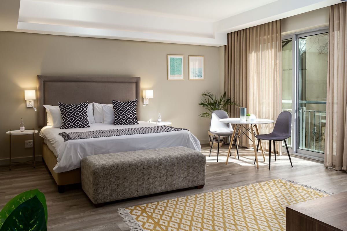 Ein Hotelzimmer mit einem großen Bett, Nachttischen, einem Schreibtisch mit Stühlen, einem Balkon mit Vorhängen und einem großen Fenster.