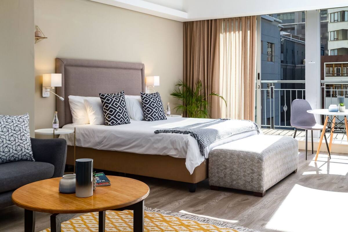 Ein modernes Hotelzimmer mit einem großen Bett, einer Sitzecke, einem kleinen Tisch und einem Balkon mit Blick auf die Stadt.