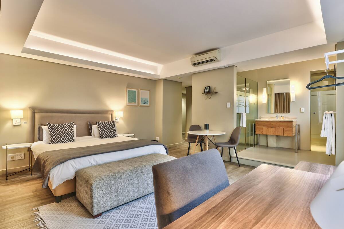 Ein modernes Hotelzimmer mit einem großen Bett, Nachttischen, einem Schreibtisch und einem durch eine Glasscheibe sichtbaren Badezimmer.
