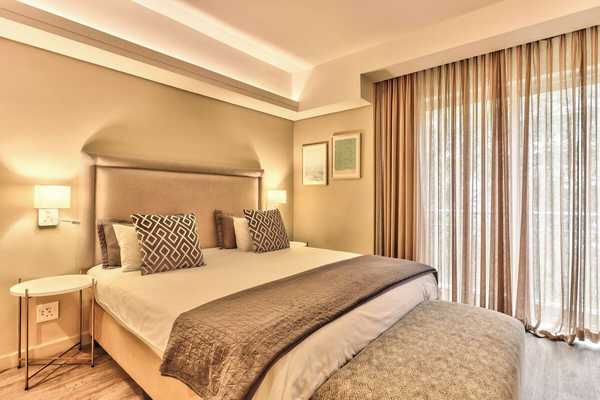 Ein Hotelzimmer mit einem großen Bett, einem Nachttisch, einer Lampe und einem Fenster mit Vorhängen.