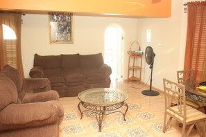 A living room with a brown sofa, a glass coffee table, a fan, and a dining area with a chair and table.