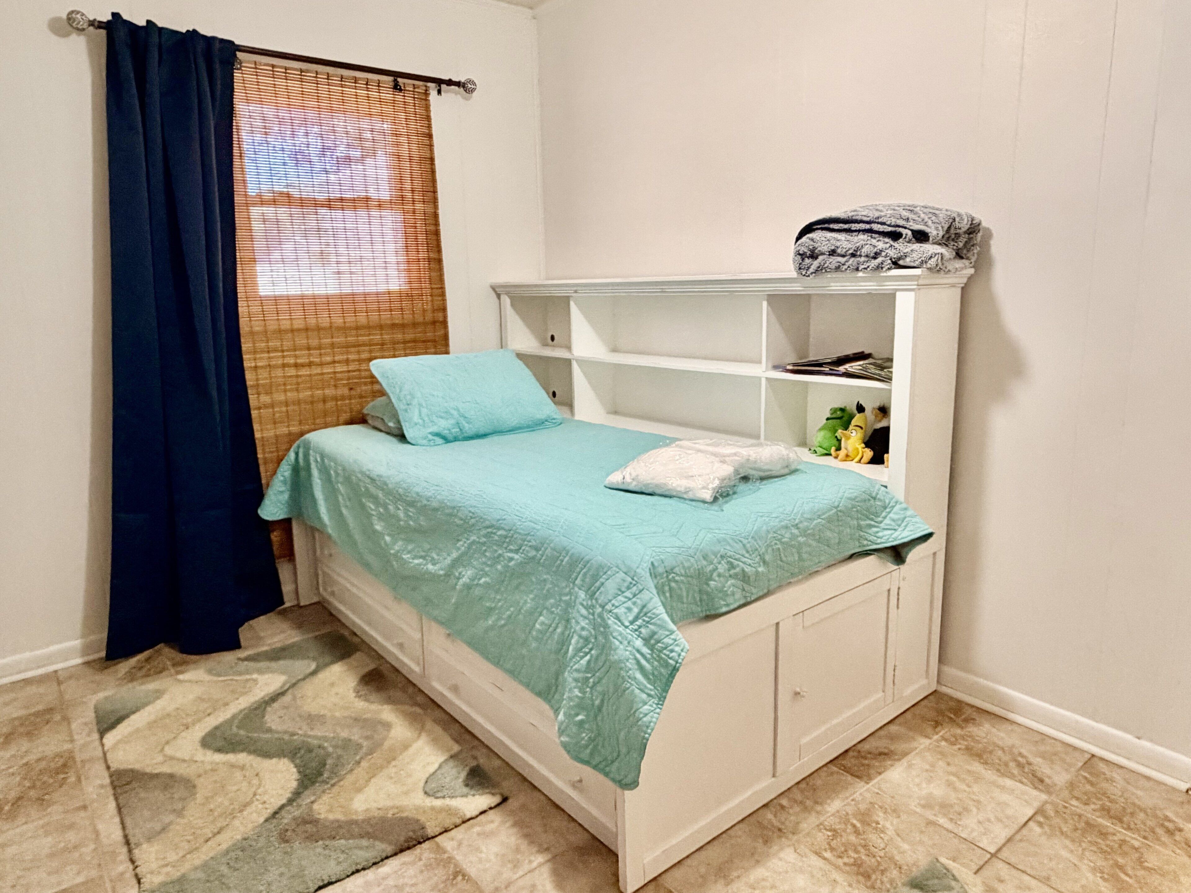 A bedroom with a bed, a white wardrobe, a shelf with items, and a window with blinds.
