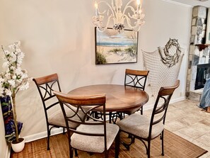 A dining area with a round table, four chairs, a chandelier, a painting of a beach scene, and a decorative wall piece.