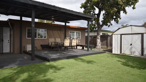 A covered patio with a table and chairs, a small lawn, and a shed in the background.