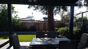 Un patio cubierto con una mesa y sillas, una terraza de madera y vistas a un jardín con césped y una cerca de madera.
