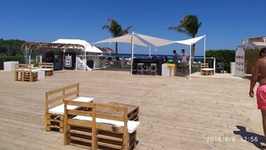 Un área frente a la playa con bancos de madera, una zona de bar con un letrero que dice "WE LOVE UP", y una vista despejada del océano.