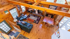 A wooden cabin interior with a loft dining area, a living room with a sofa and chairs, and a kitchen area. - Climber`s Haven- Hot Tub, Firepit, Sunroom, WiFi, 5 mins to Natural Bridge (Rogers)