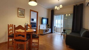 Ein Essbereich mit Holzkohlen und einem runden Tisch, ein Wohnzimmer mit einer Couch, einem Fernseher und einem großen Fenster mit Jalousien.