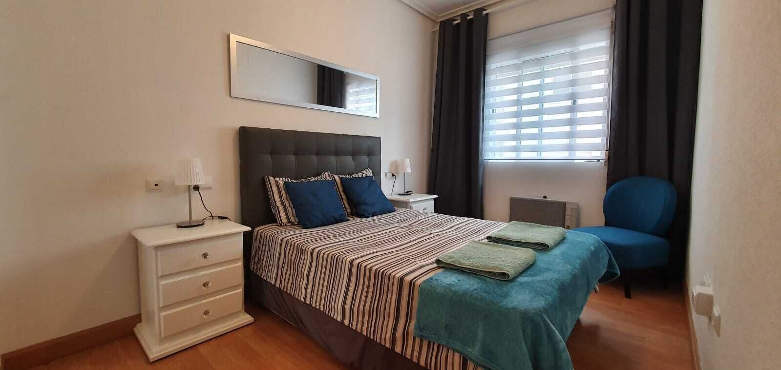 Un dormitorio con cama, mesitas de noche, una silla, un espejo y una ventana con cortinas.