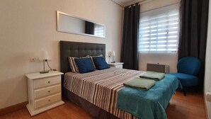 Ein Schlafzimmer mit Bett, Nachttischen, einem Sessel, einem Spiegel und einem Fenster mit Vorhängen.