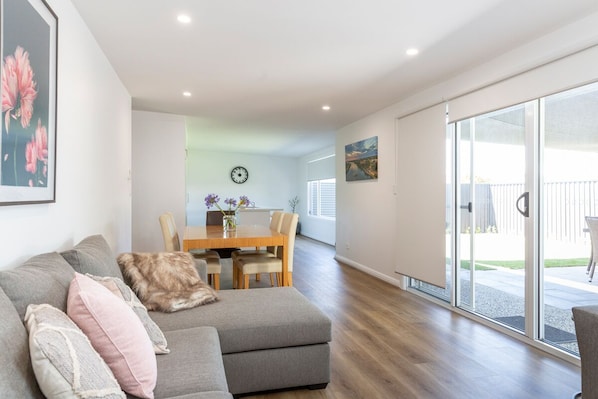 A modern living room with a grey sectional sofa, a wooden dining table with chairs, and a large sliding glass door leading to an outdoor patio.