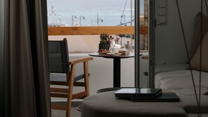 Ein Schlafzimmer mit einem Bett, einem kleinen Tisch mit Tasse und Pflanze sowie Blick auf das Meer und die Berge durch ein Fenster.