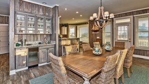 A dining room with a wooden table and wicker chairs, a kitchen with wooden cabinets, and a chandelier. - Ocean view home, remodeled with large bedrooms and living space! Wrap around porches for enjoyment (Bald Head Island)