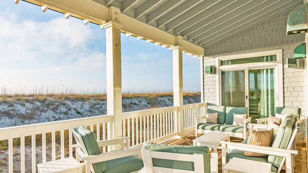 A covered outdoor seating area with white furniture and green cushions, overlooking a beach with sand dunes. - Ocean view home, remodeled with large bedrooms and living space! Wrap around porches for enjoyment (Bald Head Island)
