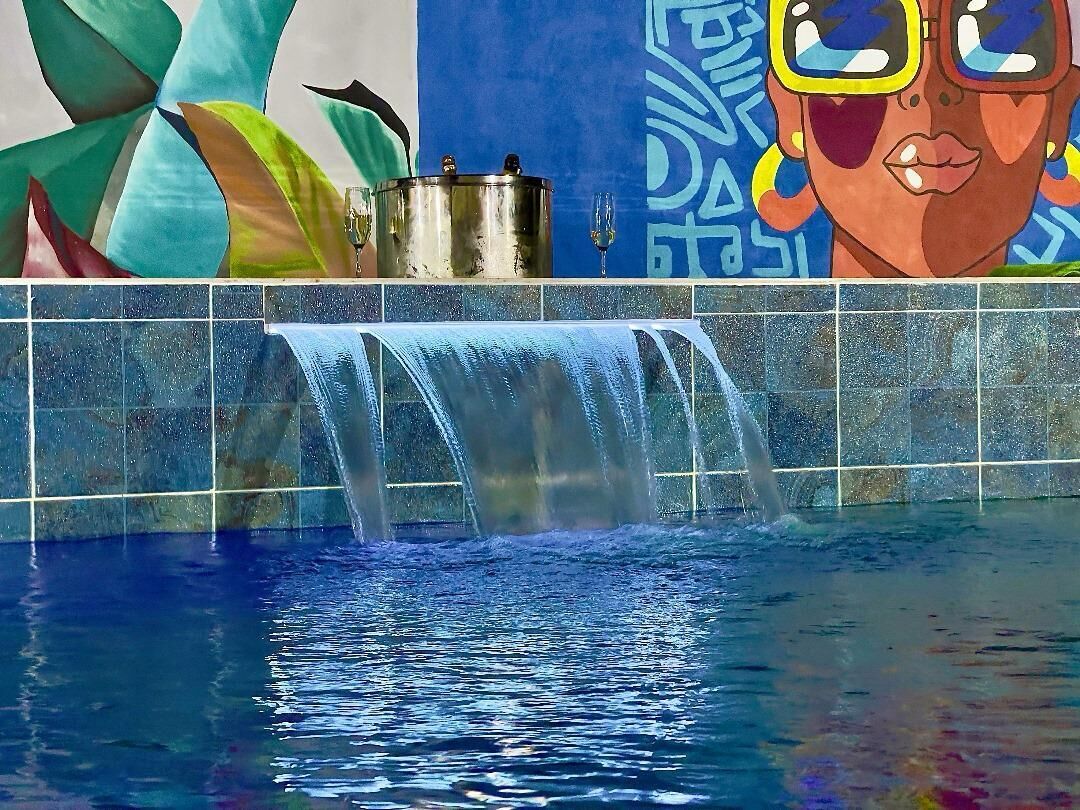 A pool with a waterfall feature, a mural with abstract art, and a metal pot on the edge.