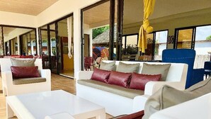 A modern outdoor seating area with white sofas and benches, large glass doors leading to an outdoor dining area. -  KIA ORA LODGE (Assinie)