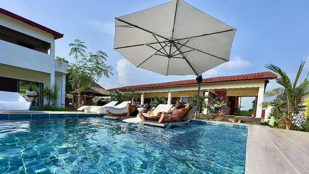 A clear swimming pool with lounge chairs and umbrellas, surrounded by buildings and palm trees. -  KIA ORA LODGE (Assinie)