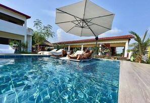 Una piscina cristalina con sillas de descanso y sombrillas, rodeada de edificios y palmeras.