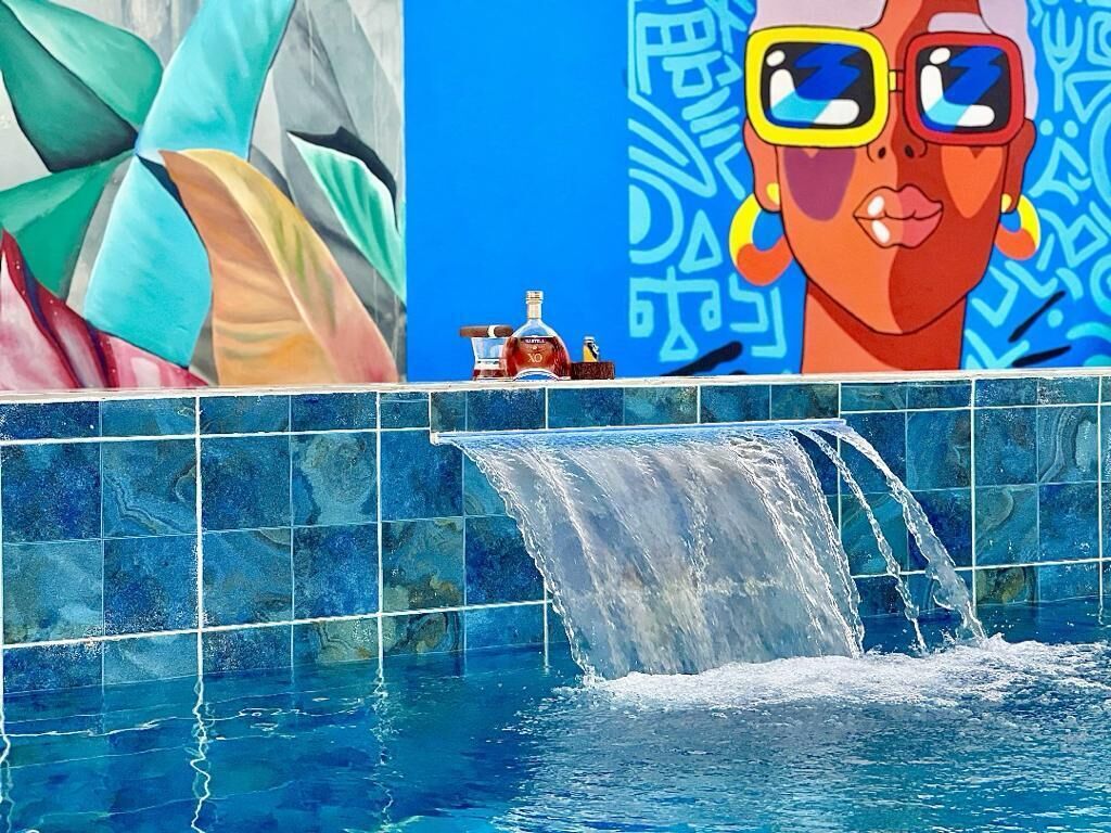 A pool with a waterfall feature, a colorful mural, and a bottle of liquor on the edge.