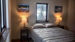 A bedroom with a bed, two bedside lamps, a window, and two paintings on the wall.