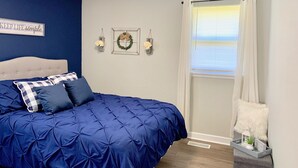 Un dormitorio con colcha azul, cabecero blanco y lámpara fijada a la pared.