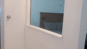 Un espacio de ducha pequeño y cerrado con ventana, puerta y una cama visible al fondo.