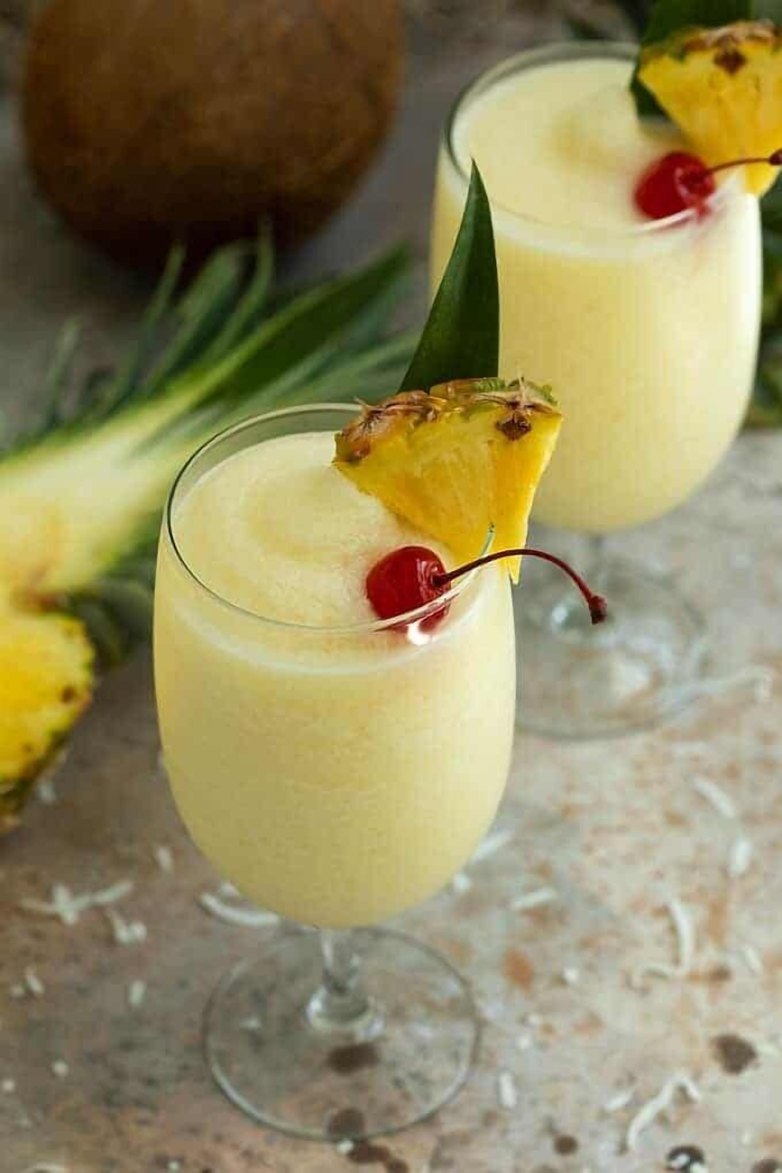 Duas taças de uma bebida amarela, decoradas com uma fatia de abacaxi e uma cereja, com um abacaxi e um coco ao fundo.