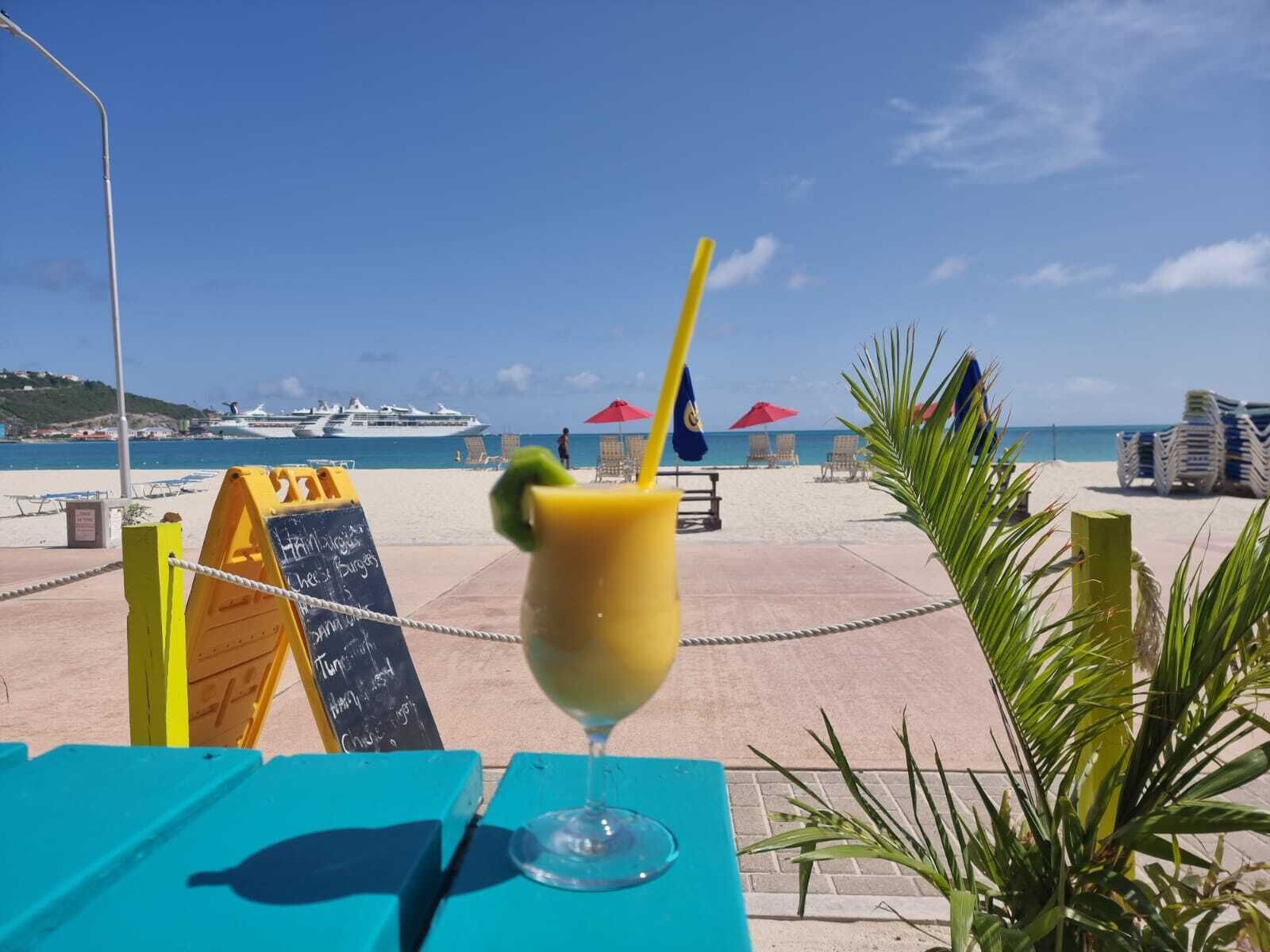Una mesa frente al mar con una bebida, un menú en pizarra y vistas al océano y cruceros.