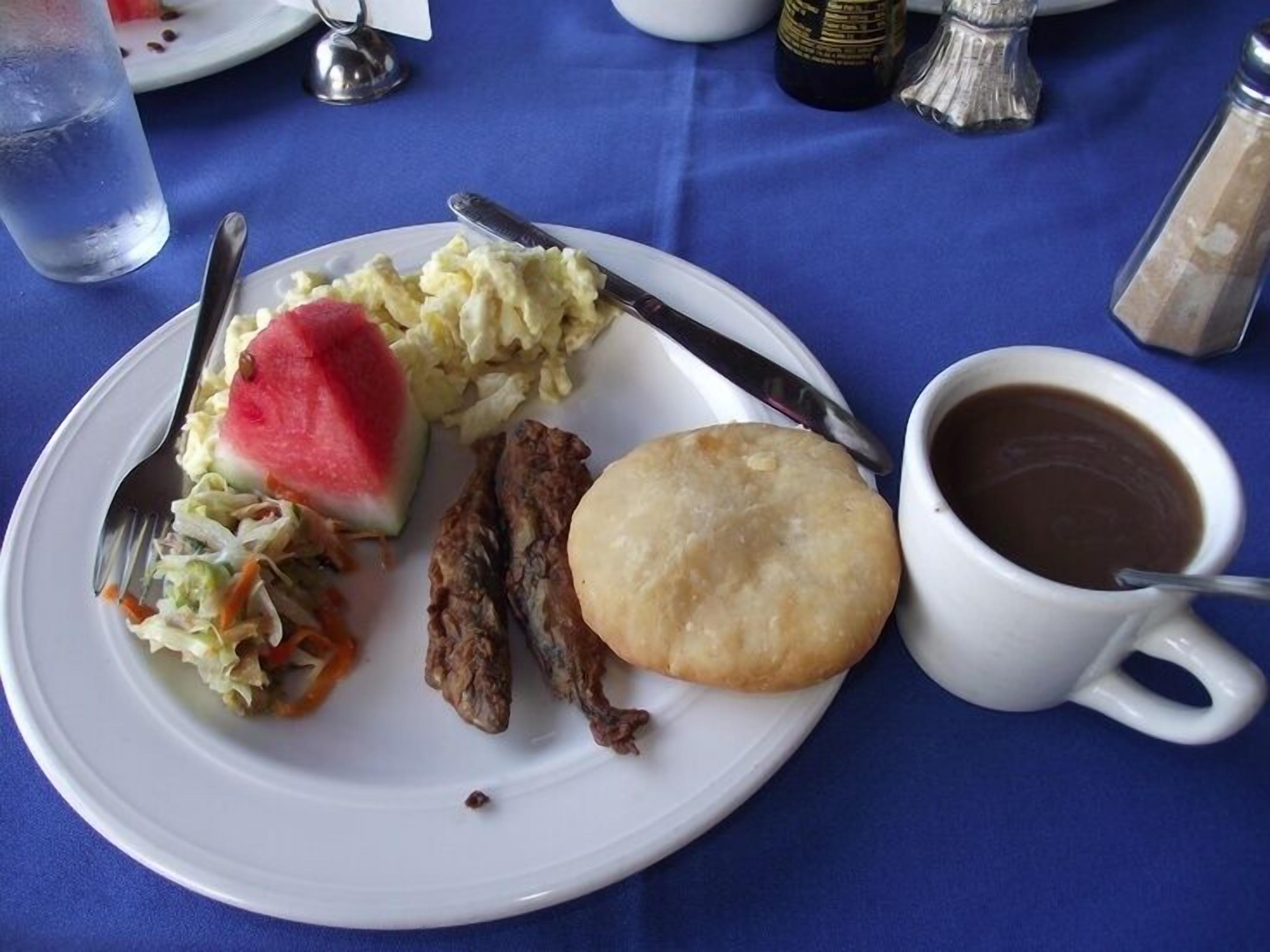 A plate with coleslaw, a slice of watermelon, a piece of meat, a biscuit, and a cup of coffee on a blue tablecloth.