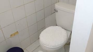 Une petite salle de bain propre, avec des carreaux blancs, des toilettes blanches, une poubelle bleue et un tapis blanc sur le sol.
