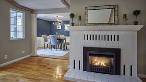 A living room with a fireplace, wooden flooring, a dining area with chairs and a table, and a large window. - Oakway Blissful Abode, Walk to Autzen Stadium (Eugene)