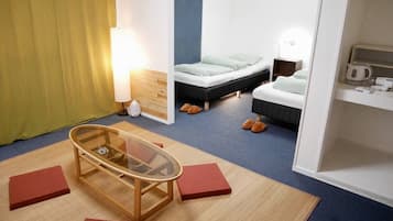Une chambre d’hôtel avec une table à plateau en verre, un lit avec une tête de lit bleue, une table de chevet avec une lampe et une petite étagère avec une bouilloire.