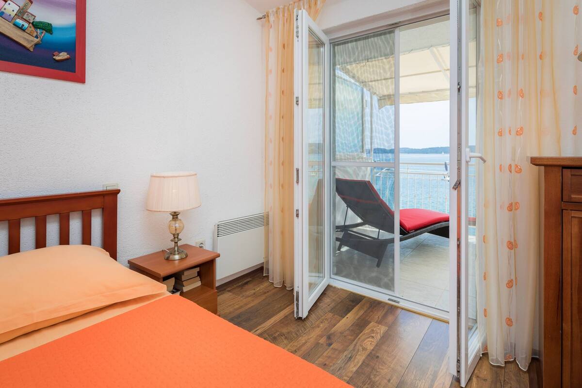 A hotel room with a bed, a nightstand with a lamp, a wooden dresser, and a sliding glass door leading to a balcony with a red chair.