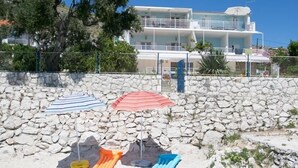 A beach with a stone wall, a tree, and a building in the background. - Gem of the Sea Alenka (Seget)