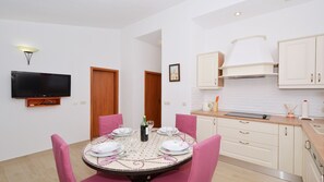 A dining area with a round table set for four, pink chairs, a flat-screen TV mounted on the wall, and a kitchen with white cabinets and a stove. - Gem of the Sea Alenka (Seget)