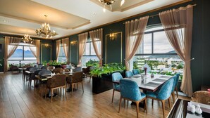A restaurant with wooden flooring, multiple dining tables, and chairs. There are large windows with curtains, and a view of a cityscape.
