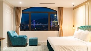 A hotel room with a large window offering a cityscape view, a bed, a blue armchair, and a matching ottoman.