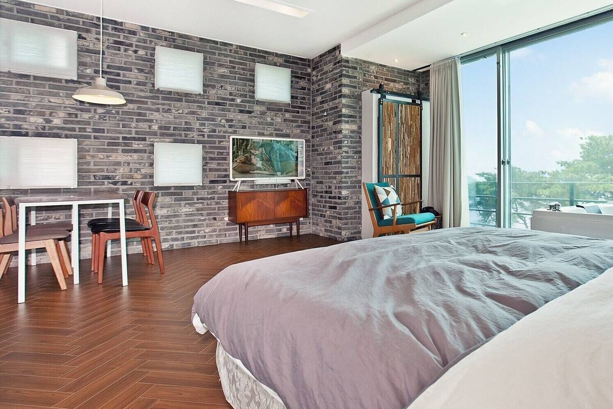 A modern bedroom with a large bed, a desk with chairs, a TV, and a large window with a view.