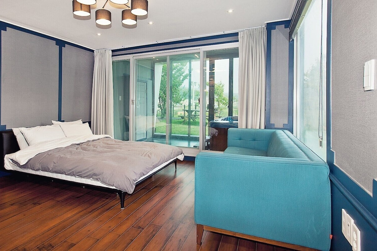 A bedroom with a bed, a blue armchair, and a large window with a view of the outdoors.