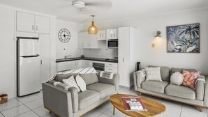 Una cocina moderna con armarios blancos, horno empotrado y microondas. Hay un sofá gris con cojines estampados y una mesa de centro de madera con revistas.