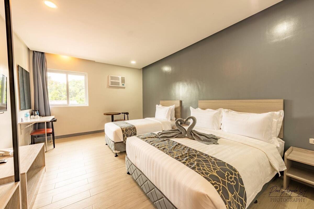 Quarto de hotel com duas camas, uma escrivaninha, uma cadeira, uma televisão e uma janela.