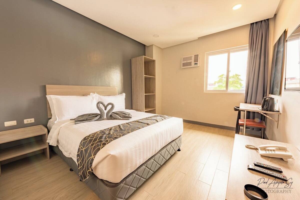 Quarto de hotel com cama, mesa de cabeceira, escrivaninha, cadeira e janela com cortinas.