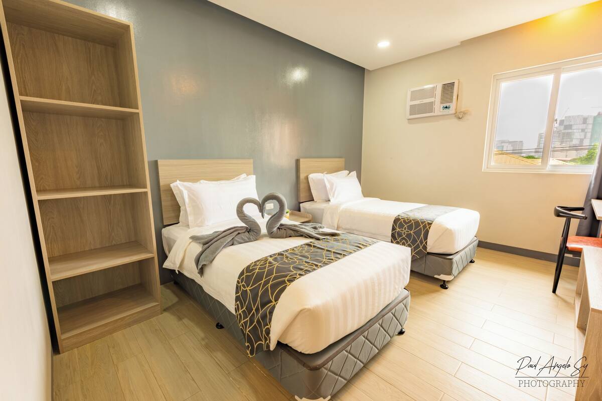 Quarto de hotel com duas camas, uma estante de madeira, uma escrivaninha e janela com vista para a cidade.