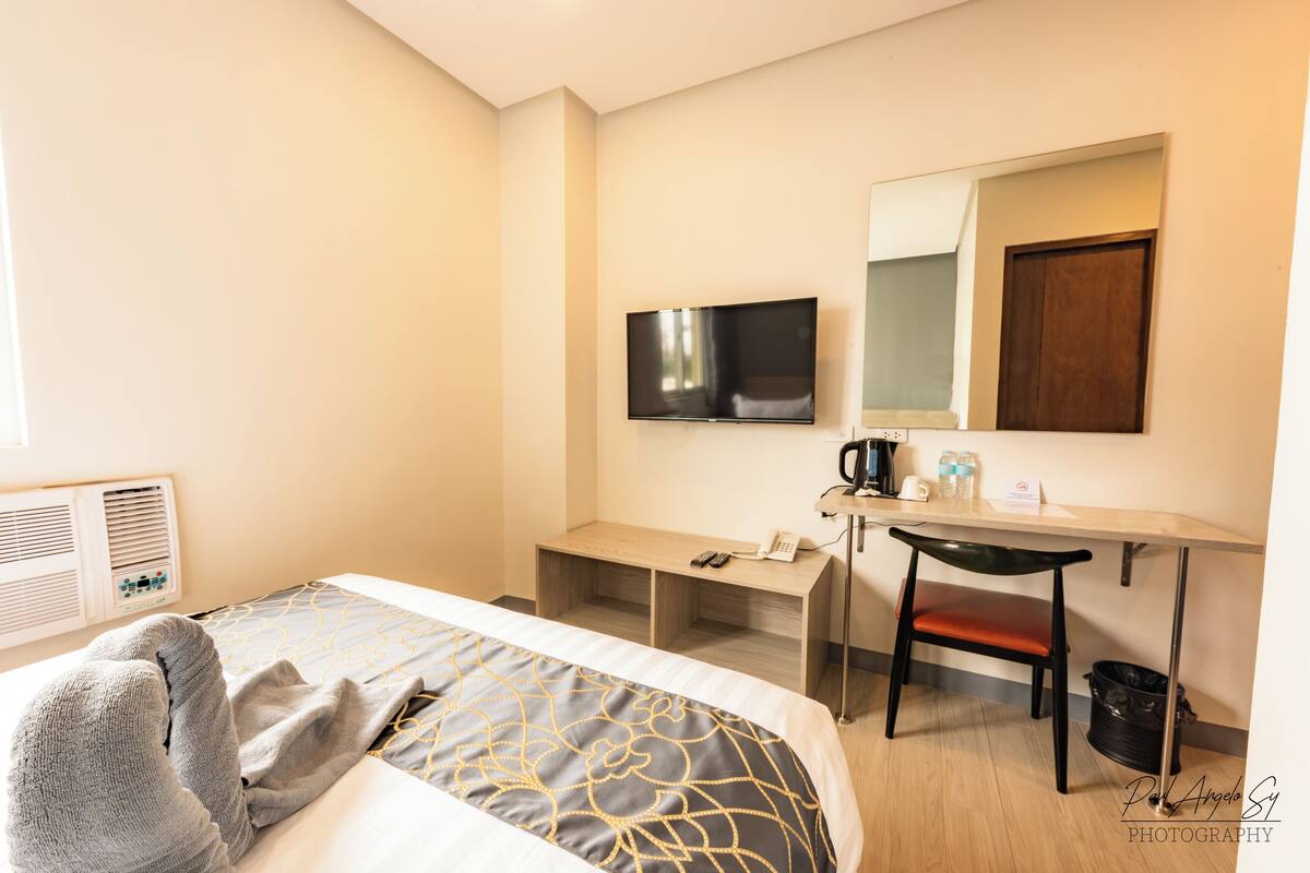 Quarto de hotel com uma cama, escrivaninha, cadeira, televisão e espelho.