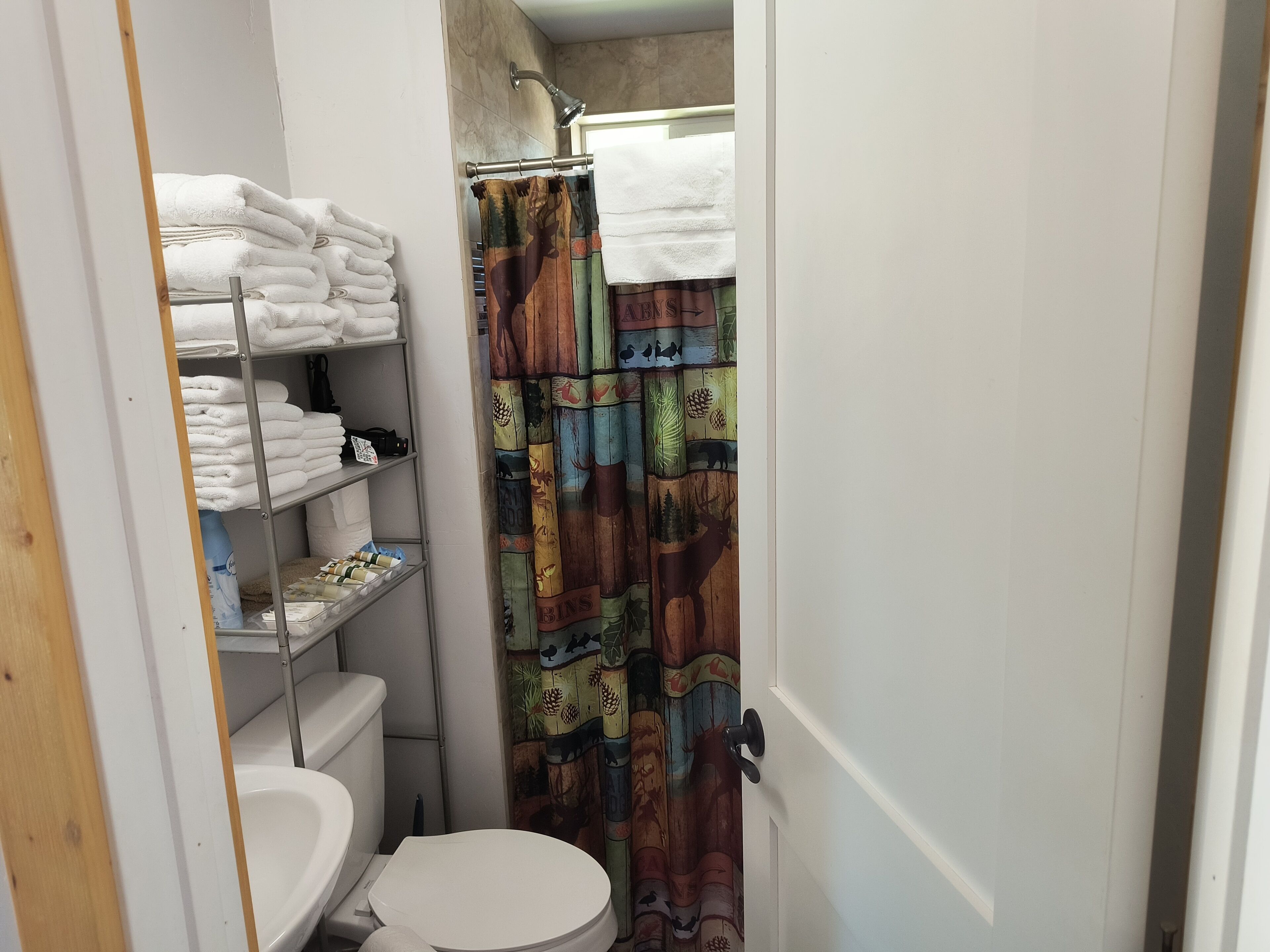 A compact bathroom with a toilet, a shelf with folded towels, and a shower curtain with a forest pattern.