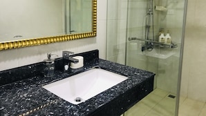 A bathroom with a white sink, black granite countertop, a gold-framed mirror, and a glass-enclosed shower area with toiletries on a shelf.
