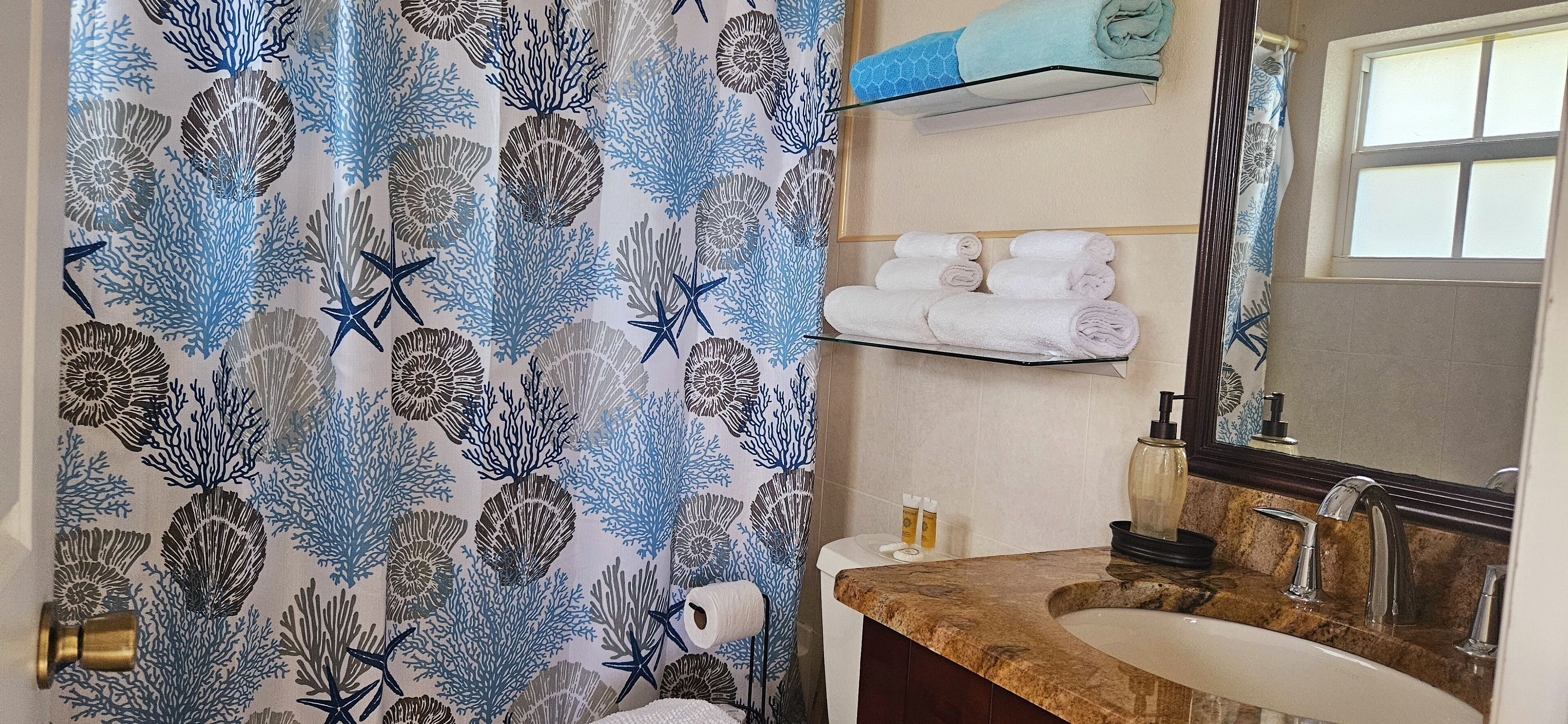 Un bagno con tenda da doccia a motivo di coralli blu e bianchi, un portasciugamani con asciugamani piegati con cura, un lavandino con piano in marmo e uno specchio sopra il lavandino.