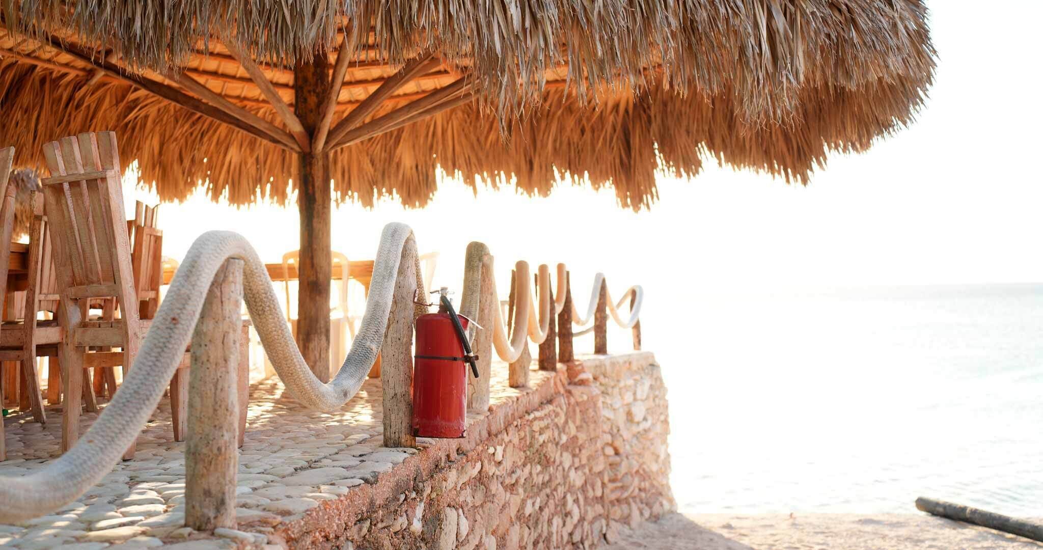 Une structure en bois située au bord de la plage, dotée d’un toit de chaume, d’un extincteur et d’une barrière en corde.
