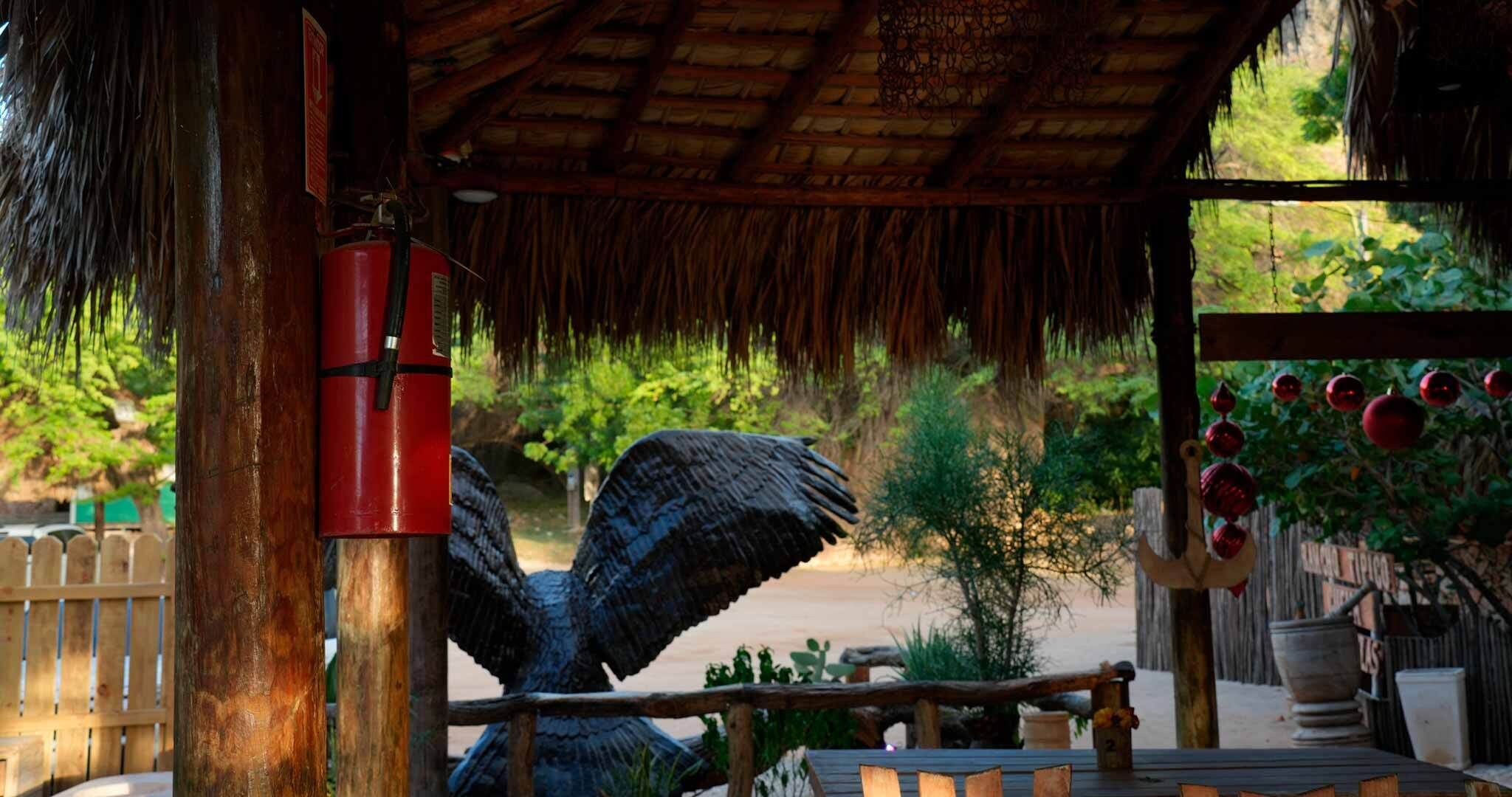 Un espace extérieur rustique doté d’une structure en bois, d’un extincteur rouge et d’une grande sculpture d’oiseau.